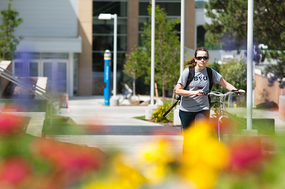Katherine Ziemann, walking her bike on campus in the Central Quad between the Manwaring center and the McKay Building with BYU-Idaho center behind her and colorful flowers in the foreground.