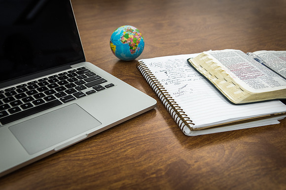 A computer, notebook and scriptures sit on a desk.