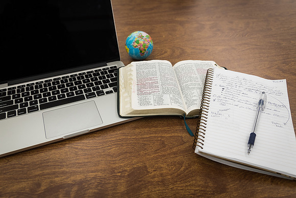 A computer, notebook and scriptures sit on a desk.
