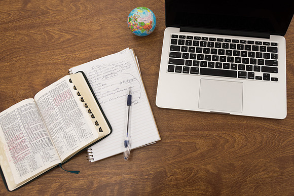 A computer, notebook and scriptures sit on a desk.