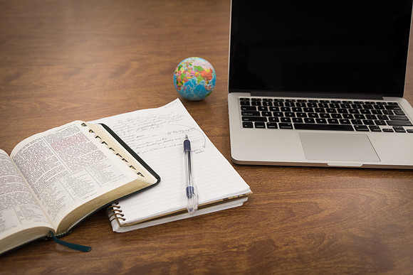 A computer, notebook and scriptures sit on a desk.