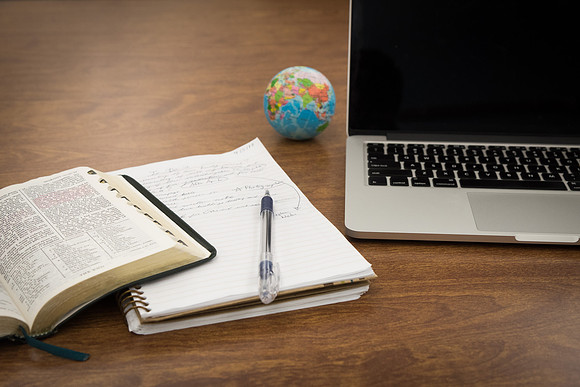 A computer, notebook and scriptures sit on a desk.